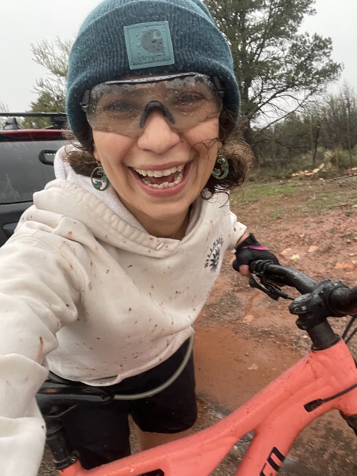 Anya Metcalfe grinning on a mountain bike, spattered with red mud on a rainy trail.