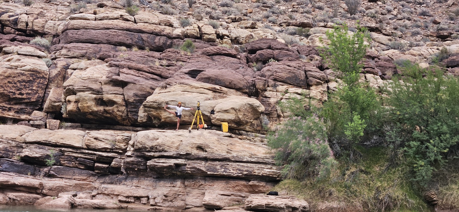 Anya Metcalfe balancing on one leg beside a surveying tripod on a sandstone ledge above the Colorado River, dwarfed by the canyon wall behind her.