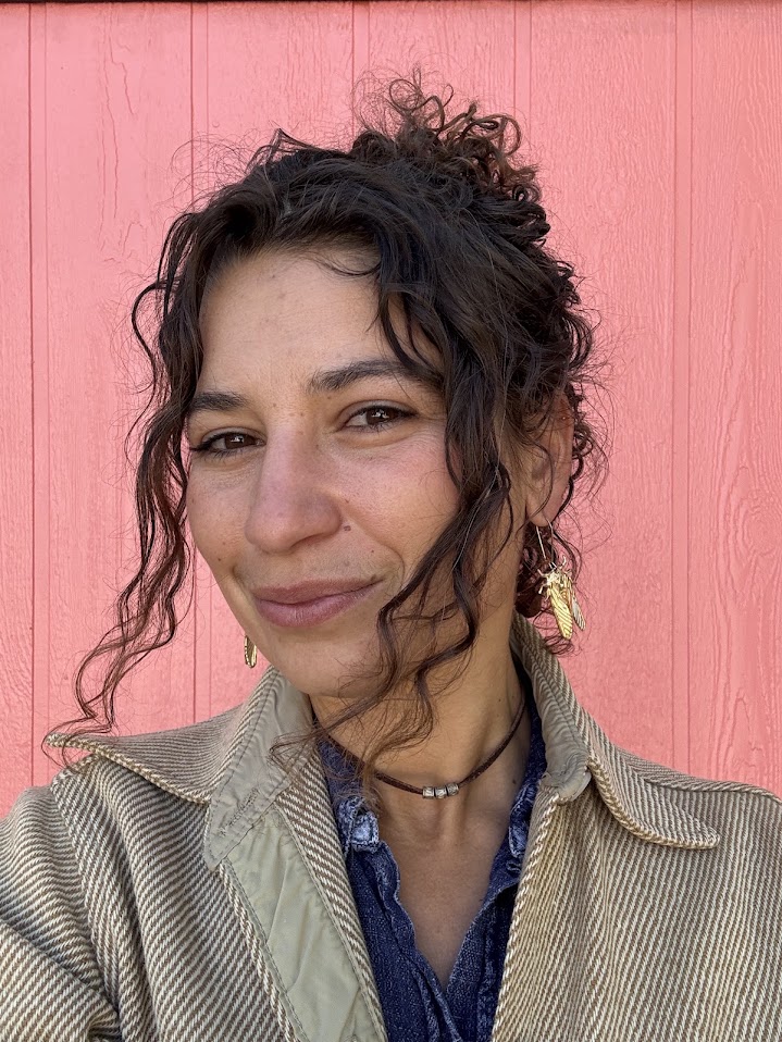 Anya Nova Metcalfe smiling, wearing a cicada earring, against a coral-painted wall.