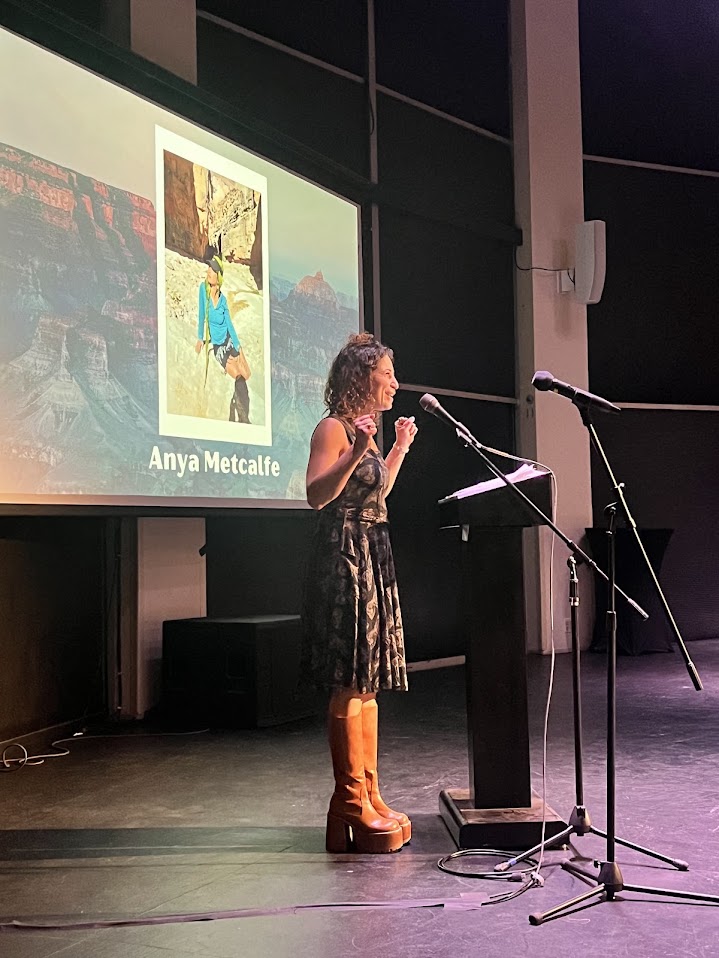 Anya Metcalfe speaking at a microphone on a stage, in front of a projected slide.