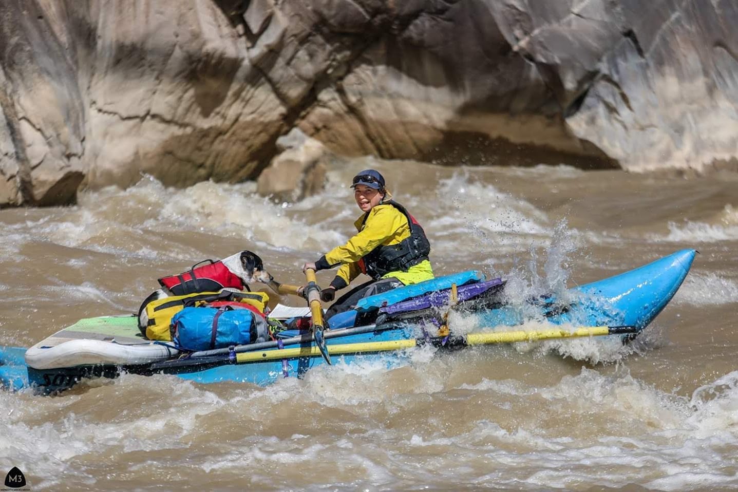 Anya Metcalfe rowing a loaded cataraft through whitewater in a canyon, with a dog in a life jacket riding on the front tube.